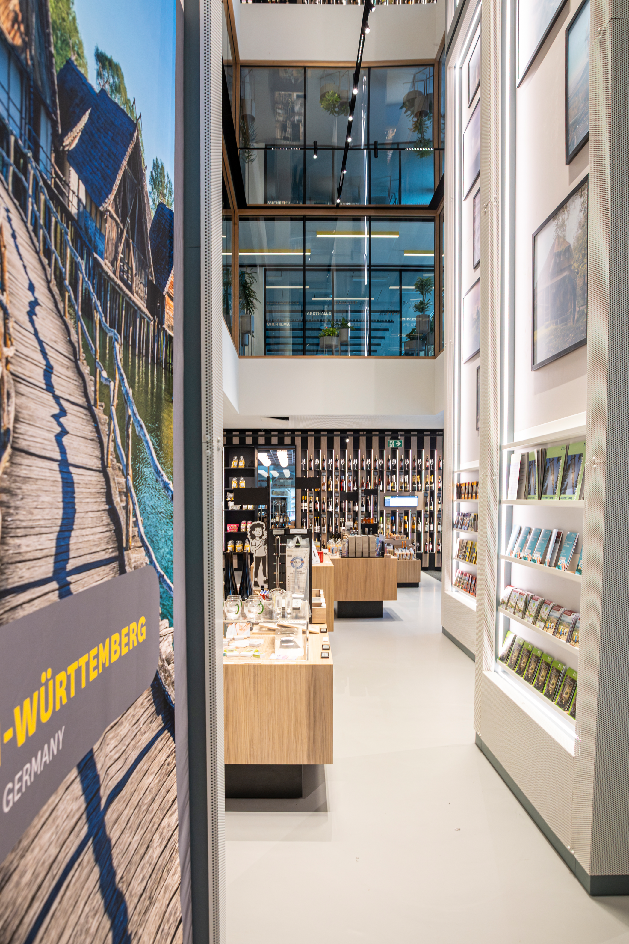 i-Punkt Verkaufsraum, Stuttgart-Marketing GmbH, Sarah Schmid Blick in die Tourist-Information, Richtung Wein-Wand. Auslage von Produkten und Broschüren auf Tischen und in Regalen. Durch die hohe Decke ist ein Blick in die Ebene 1, wo sich die Tagungsräume befinden, möglich.