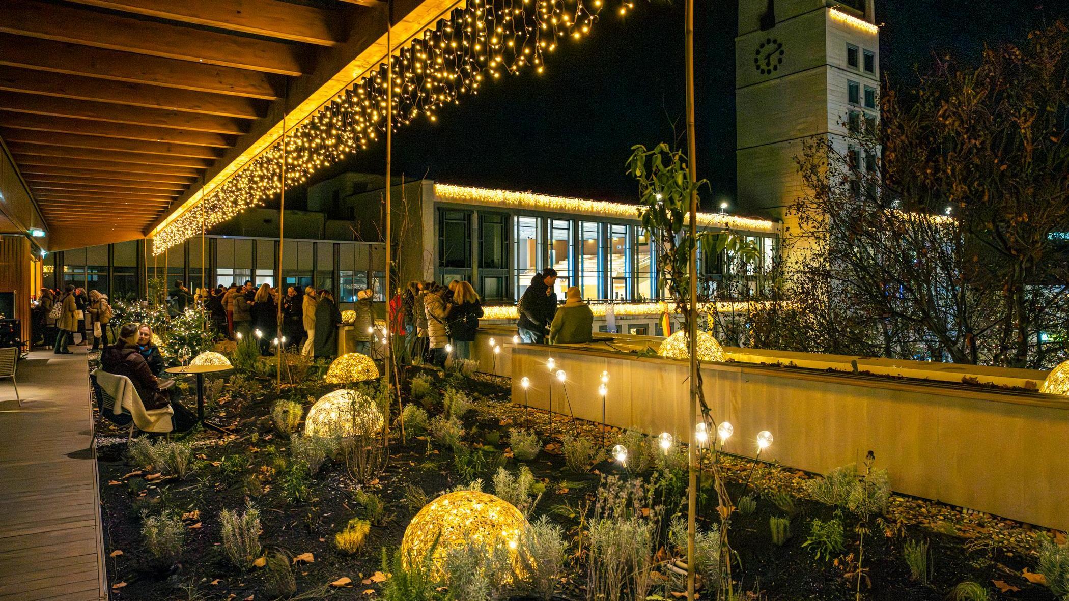 Dachterrasse zur Weihnachtszeit, Dachterrasse des Haus des Tourismus mit vielen Lichtern: beleuchtete geflochtene Halbkugeln im Blumenbeet, Lichterkette am Dach und Leuchtkugeln auf Stäben. Im Hintergrund stehen Personen in warmer Kleidung zusammen.