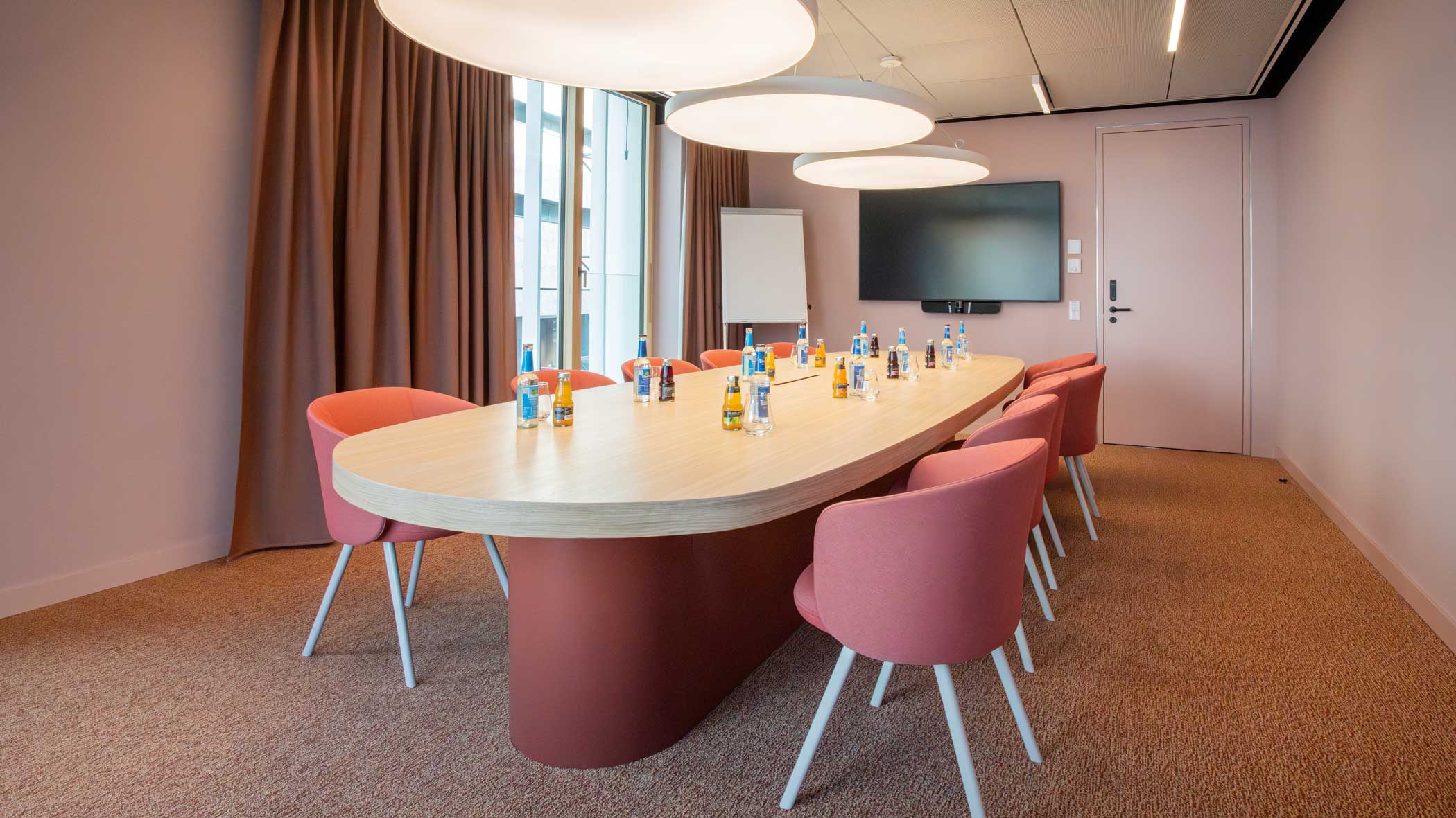 Rote Stühle angeordnet um einen ovalen Tisch; an der Stirnseite des Raums sind ein großer Bildschirm und ein Flipchart.