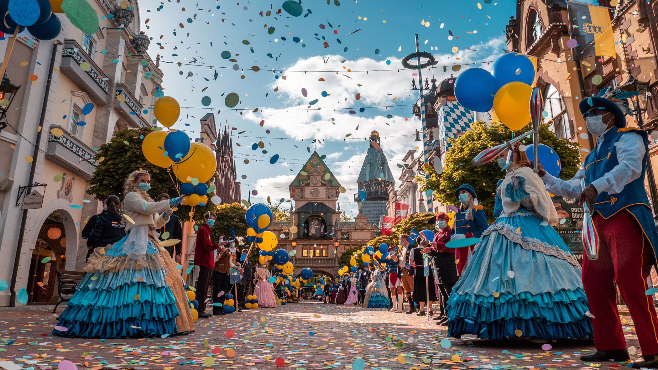 Kostümierte Personen stehen Spalier zu einem großen Portal. Bunte Kleider und Anzüge, gelbe und blaue Luftballons sowie buntes Konfetti dominieren das Bild. 