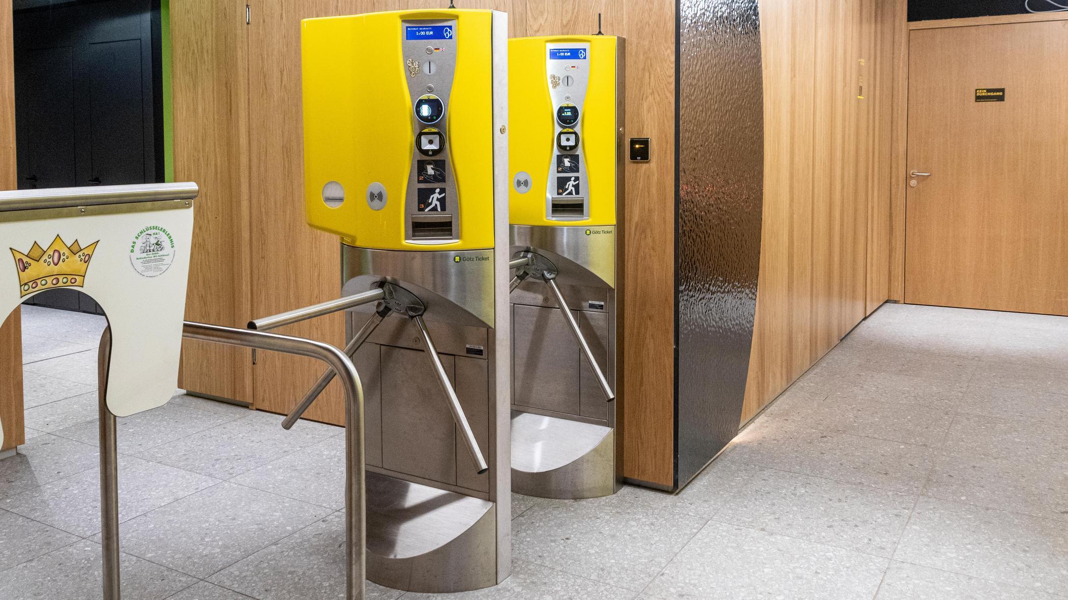 Zwei Drehkreuze markieren den Zugang zu den Toiletten mit zwei gelben Ticketautomaten. Links davon der Zugang für Kinder, Kinderwägen, Rollstühle und Gehhilfen.