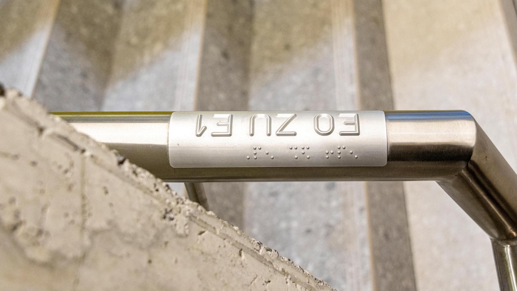 Ein Handlauf mit taktiler Beschriftung und Brailleschrift zur Orientierung für Seheingeschränkte in einem Treppenhaus.