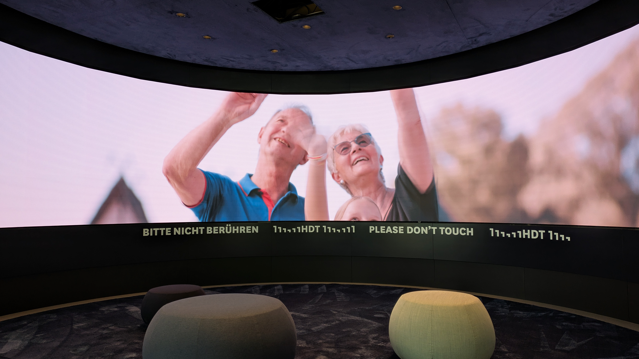 Ausschnitt der großen LED-Anzeige, die im Halbkreis aufgestellt ist. Das Video zeigt drei Personen, die einem Zeppelin nachblicken. Der Boden des Vorführbereichs ist mit Teppich ausgelegt. Die Decke ist ebenfalls mit Stoff überzogen. Drei Sitzgelegenheiten laden zum Verweilen ein.