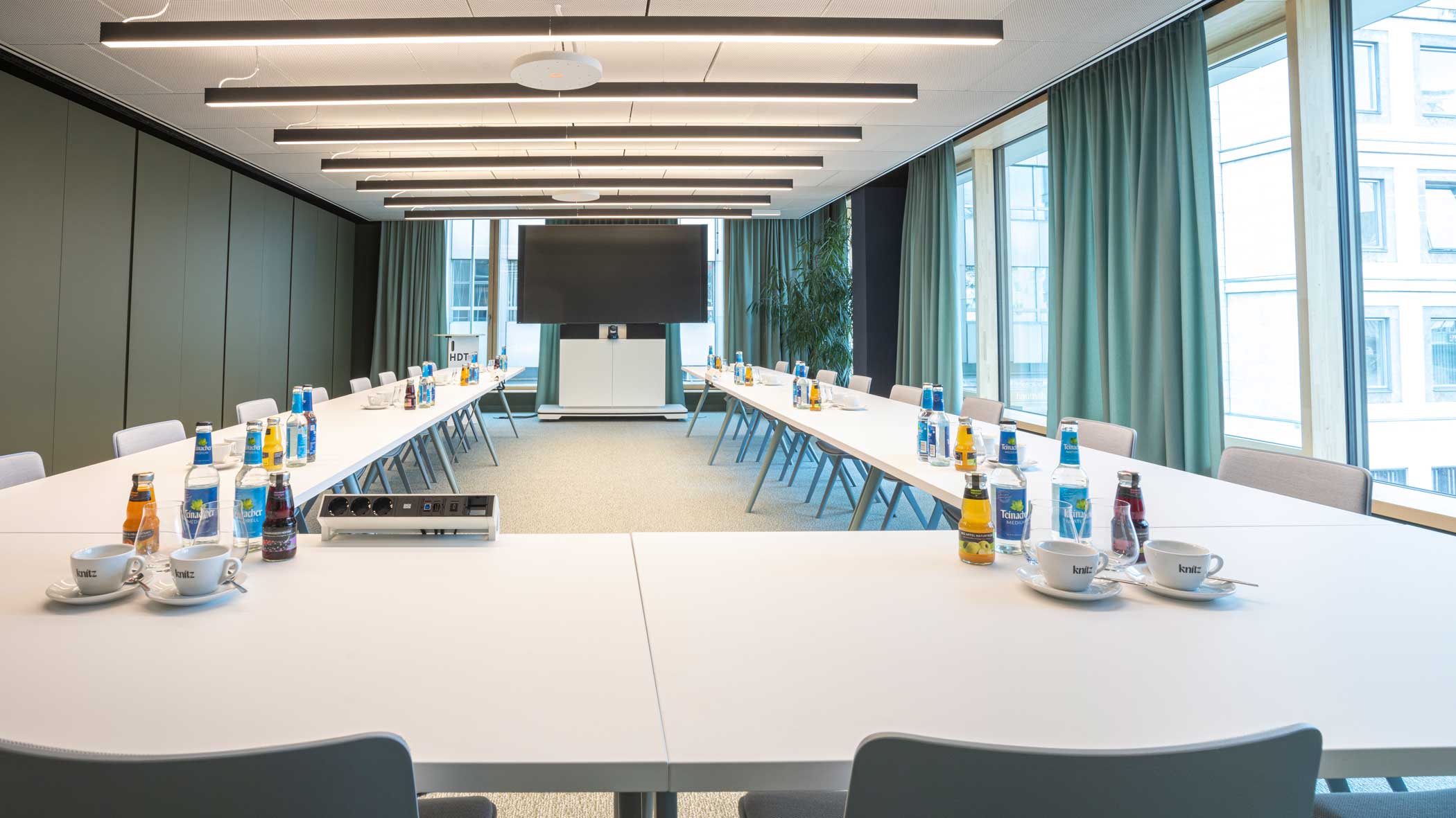 Klassischer Konferenzraum in gedeckten Farben mit U-förmig gestellten weißen Tischen und Bestuhlung. An der Stirnseite befindet sich ein großes Bildschirm.